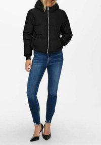 Sort pufferjakke med hætte, der har en frontlynlås, ribbede manchetter og en quiltet design. Sammen med blå skinny jeans og sorte hæle.