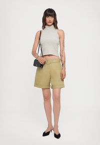Khakigrüne Shorts mit Gürtel und Seitentaschen, kombiniert mit einem eng anliegenden gestreiften Rollkragenpullover und einer kleinen schwarzen Umhängetasche. Schwarze spitze Schuhe.