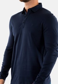 Polo a maniche lunghe di colore navy, realizzato in cotone liscio, con colletto a bottoni e un piccolo logo sul lato sinistro del petto. Design morbido e aderente.