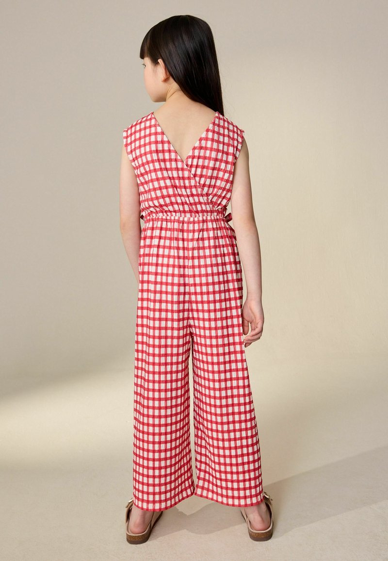 Ung pige med langt mørkt hår iført rød- og hvidternet ærmeløs jumpsuit, stående barfodet på en beige gulv mod en ensfarvet væg.