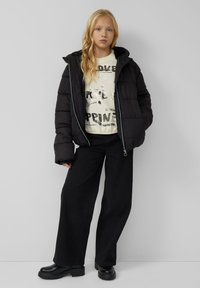 Ragazza bionda che indossa una giacca a vento nera, una felpa con grafica color crema, pantaloni larghi neri e stivali chunky neri, in piedi contro un muro grigio.