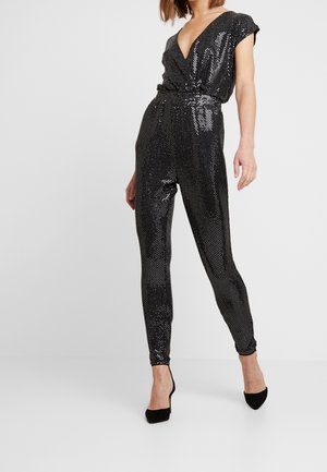 Frau trägt einen schwarzen, mit Pailletten besetzten V-Ausschnitt-Jumpsuit mit kurzen Ärmeln und schwarzen spitz zulaufenden High Heels, steht vor einem einfarbigen weißen Hintergrund.