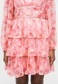 Γυναίκα με ροζ διάτρητο στρωματοποιημένο φόρεμα με μακριά διάφανα μανίκια και πλισέ ζώνη στη μέση, με τα χέρια ακουμπισμένα στο πλάι.