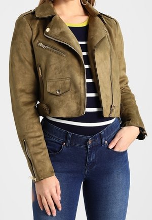 Vrouw draagt een olijfgroene, korte suède motorjas over een zwart-wit gestreepte top met blauwe jeans, één hand in de zak.