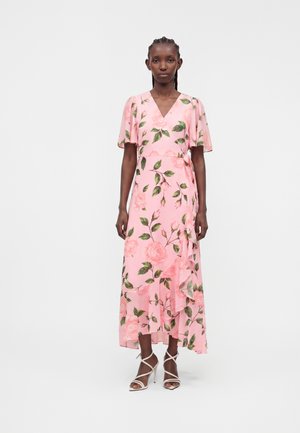 Frau steht und trägt ein pinkes Wickelkleid mit grünen Blatt- und Rosenmustern, flatternden Ärmeln und weißen, hochhackigen Riemchensandalen.