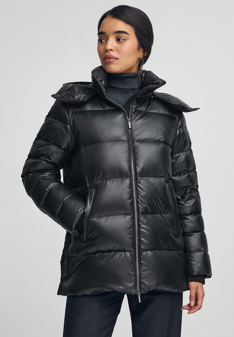 Svart puffjacka med en glänsande, quiltad design. Har dragkedja fram, hög krage och två sidofickor. Tyget ser mjukt och lent ut.