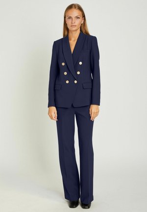 Kvinde iført en marineblå dobbeltradet blazer med guldknapper og matchende bukser, stående mod en ensfarvet lys baggrund.