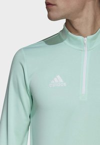 Helles mintgrünes Sporttop mit langen Ärmeln, halbem Reißverschluss und weißem Adidas-Logo auf der linken Brust. Glattes, dehnbares Material.