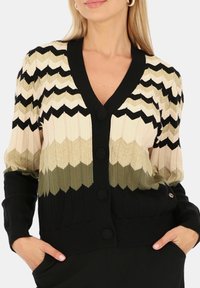 Sweter z dzianiny z dekoltem w serek, posiadający wzór w zygzak w kolorach kremowym, złotym, oliwkowym i czarnym. Pięć czarnych guzików i ściągacze przy mankietach.