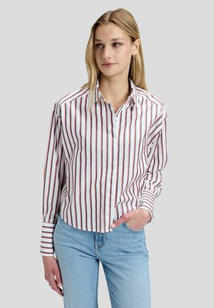 Donna che indossa una camicia bianca a maniche lunghe con righe verticali rosse e nere e jeans azzurri, in piedi davanti a uno sfondo grigio chiaro e uniforme.