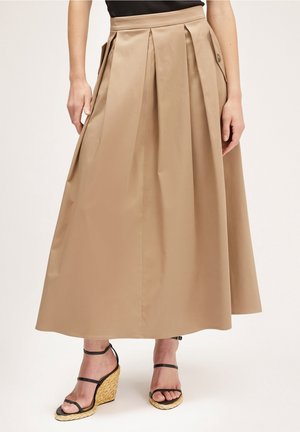 Kvinde iført en beige A-formet midi nederdel med forreste folder og sidelommer, kombineret med sorte kilehælssandaler med stropper.