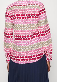 Blus med lång ärm i rosa, röd och beige cirkulära mönster. Lättviktsmaterial med rundad nederkant och knäppning vid ärmsluten. Bakifrån sedd.