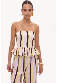 Femme portant un top bustier à volants et un pantalon assorti à rayures verticales rose, jaune, blanc et marron, accessoirisé avec des boucles d’oreilles et un bracelet en or.