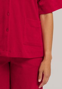Camisa roja de manga corta con botones en el frente, que cuenta con un bolsillo de parche. La tela se ve suave con una textura liviana, acompañada de pantalones a juego.
