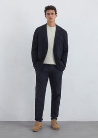 Navy blazer over a cream knit sweater, paired with navy trousers and tan suede shoes. The blazer features two front pockets and a single button.