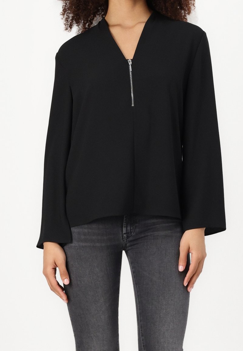 Femme portant un chemisier noir à manches longues zippé, associé à un jean gris foncé, debout devant un fond blanc uni.