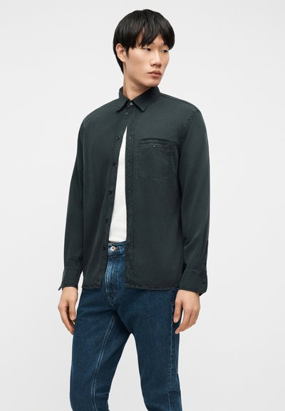 Młody mężczyzna w ciemnozielonej koszuli na guziki, nałożonej na białą koszulkę, oraz w niebieskich dżinsach, stoi na tle jasnego, jednolitego tła.