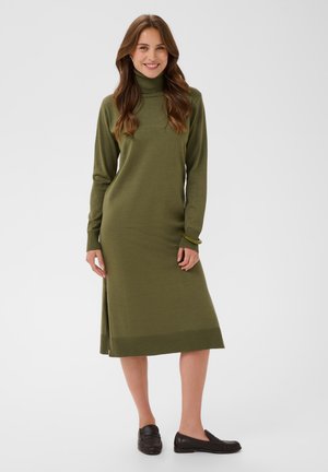 Femme souriante, portant une robe pull à col roulé vert olive à manches longues et des mocassins noirs, debout devant un fond clair uni.