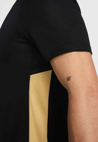 Schwarzes Kurzarmhemd mit goldener Seitenbahn; glatter Stoff und figurbetontes Design. Sichtbares Tattoo am Arm mit fünf horizontalen Linien.