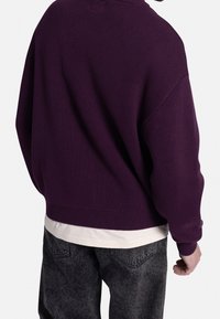 Lilla strikket sweater med løs pasform, ribbede manchetter og kant. Båret over en hvid skjorte, kombineret med mørkegrå teksturerede bukser.