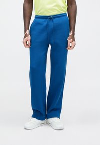 Μπλε φόρμες από απαλό, υφασμάτινο ύφασμα με κορδόνι στη μέση και στενόμακρα πόδια, συνδυασμένες με λευκά αθλητικά παπούτσια.