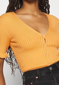 Pomarańczowy prążkowany sweterek z głębokim dekoltem w kształcie litery V i krótkimi falbaniastymi rękawami, z guzikami z przodu i detalem w kształcie dziurki. W połączeniu z czarnymi dżinsami.