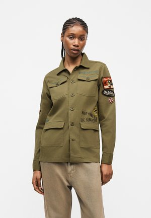 Nainen, jolla on letitetyt hiukset, pukeutuneena oliivinvihreään nappiin painettuun takkiin, jossa on peitoja ja tekstiä, yhdistettynä beigen värisiin housuihin yksiväristä taustaa vasten.