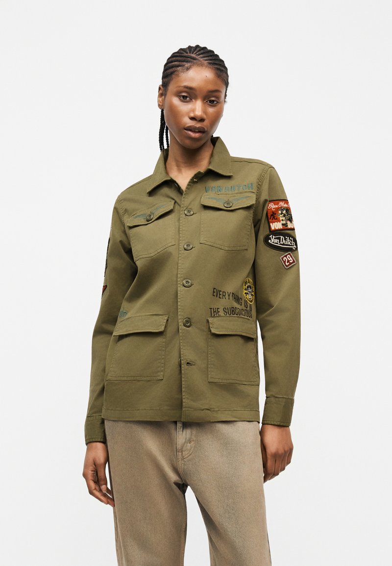 Γυναίκα με πλεξούδες που φοράει χιτώνα ελιάς με κουμπιά και μπαλώματα και κείμενο, συνδυασμένη με μπεζ παντελόνι μπροστά σε μονόχρωμο φόντο.