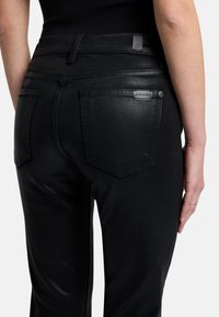 Czarne dżinsy o dopasowanym kroju, z tylnymi kieszeniami i błyszczącą fakturą. Zawierają małą metkę marki w okolicy pasa.