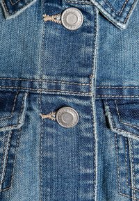 Jeansjacka fram med två silverknappar, blå färg, ljus blekning, kontrasterande sömmar och en fickdetalj på vänster sida.