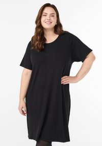 Pitkät aaltoilevat hiukset omaava nainen, joka pukeutuu löysään mustaan lyhythihaseen mekkoon, hymyilee ja pitää toista kättään lanteillaan valkoista taustaa vasten.