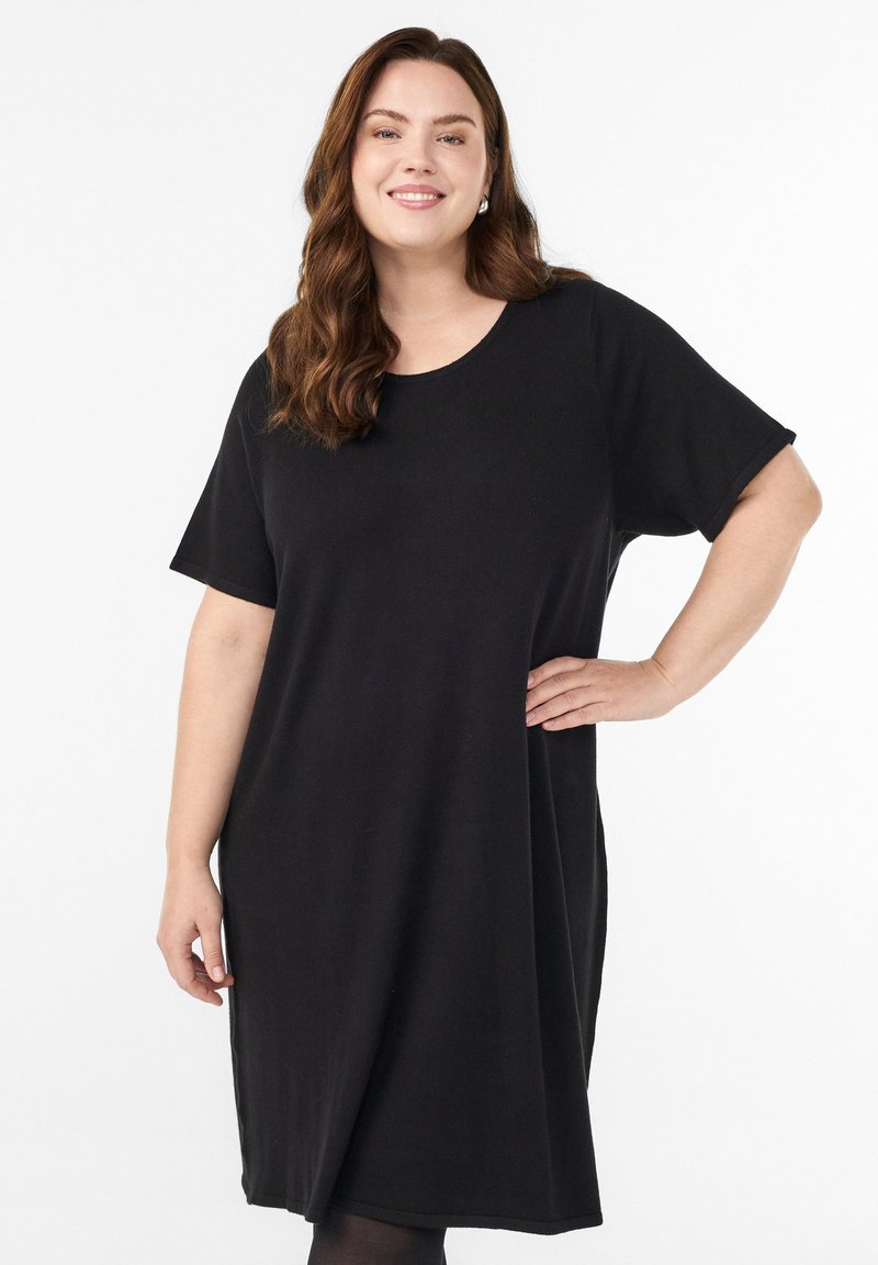 Pitkät aaltoilevat hiukset omaava nainen, joka pukeutuu löysään mustaan lyhythihaseen mekkoon, hymyilee ja pitää toista kättään lanteillaan valkoista taustaa vasten.