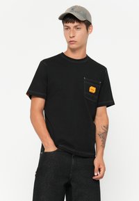 Μαύρο κοντομάνικο μπλουζάκι με τσέπη στο μπροστινό μέρος και πορτοκαλί ετικέτα. Φτιαγμένο από βαμβάκι με αντίθετη λευκή ραφή. Φοράει μαύρα τζιν.