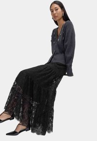 Grå cardigan med texturerad stickning och dekorativa detaljer, kombinerad med en lång, flödande svart spetskjol med lager på lager av volanger. Svarta skor.