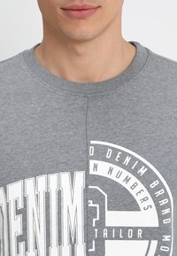 Sudadera gris con un cuello redondeado de canalé, que presenta un audaz texto gráfico en blanco y un diseño de logo circular en el pecho. Textura suave.