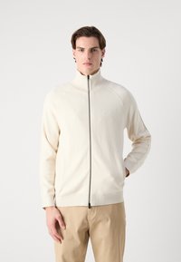 Ljusbeige zip-up-tröja med hög krage, mjuk textur och raglanärmar. Har en synlig svart dragkedja framtill.