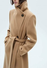 Dlouhý béžový vlněný kabát s vysokým límcem, dvouřadým zapínáním, páskem v pase a velkými černými knoflíky.