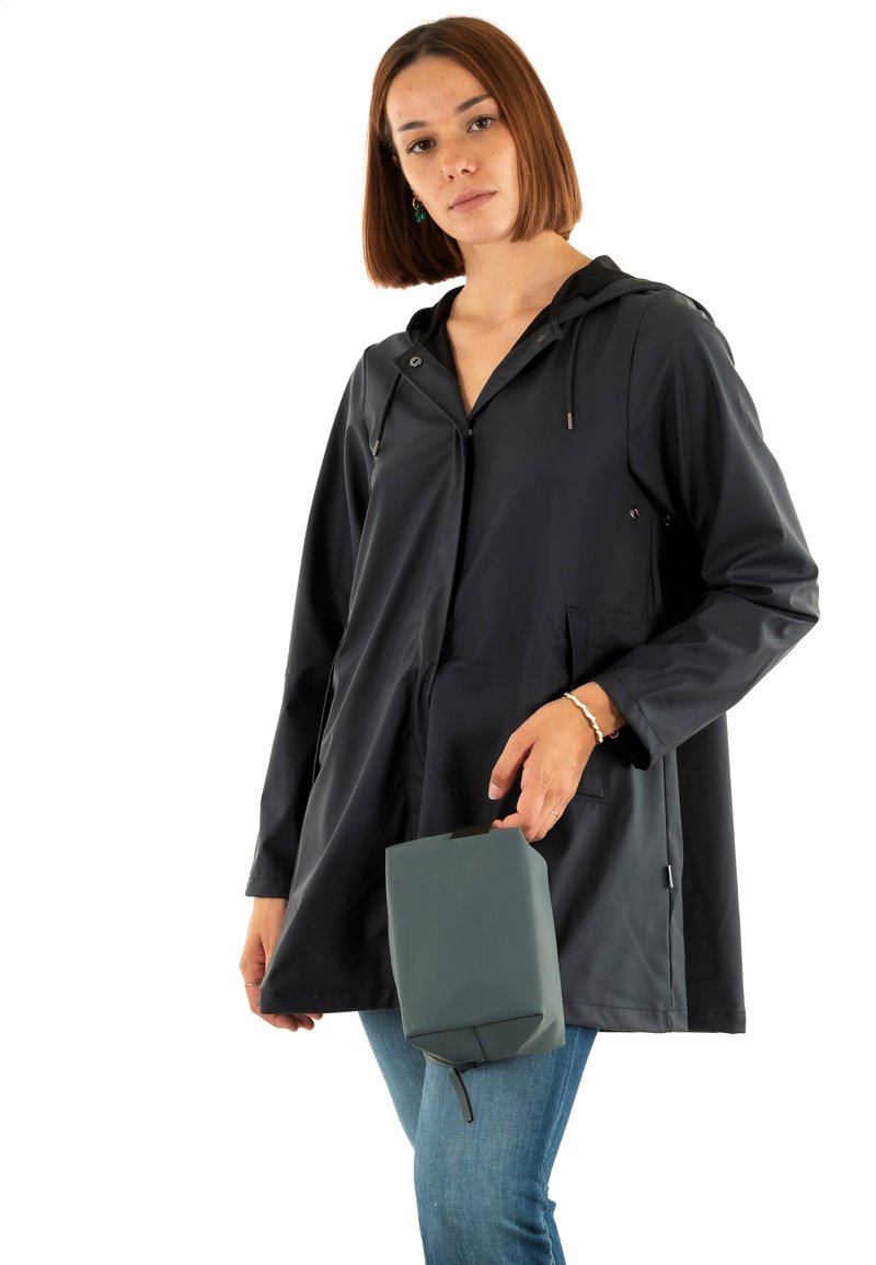 Cappotto impermeabile nero con cappuccio, maniche lunghe e tasche frontali; abbinato a una borsa rettangolare grigia, indossato con jeans blu.
