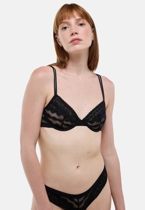 Soutien-gorge en dentelle noire avec panneaux transparents, bretelles fines réglables et bordure festonnée. Associé à un string en dentelle assorti. Texture lisse.