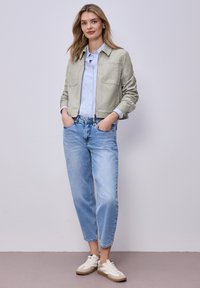 Mujer joven de pie con las manos en los bolsillos, vistiendo una chaqueta beige, camisa azul claro, vaqueros cortos azules y zapatillas blancas, sobre un fondo liso.