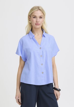 Ženska z dolgimi blond lasmi nosi svetlo modro kratko srajco z gumbi, temne hlače, zlate uhančke in črno zapestnico na desnem zapestju.