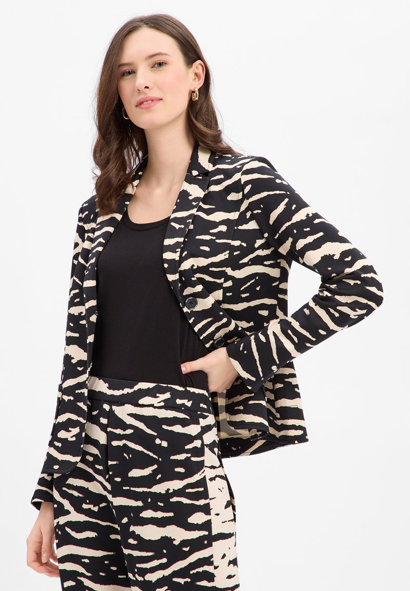 Zwart- en crèmekleurige blazer met een patroon, een getailleerde pasvorm, knoopdetails en lange mouwen, gecombineerd met een zwarte top en een broek met een vergelijkbaar patroon.