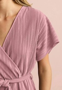 Primer plano de una persona que lleva un vestido envolvente de color malva claro, con mangas cortas y un cinturón atado a la cintura.