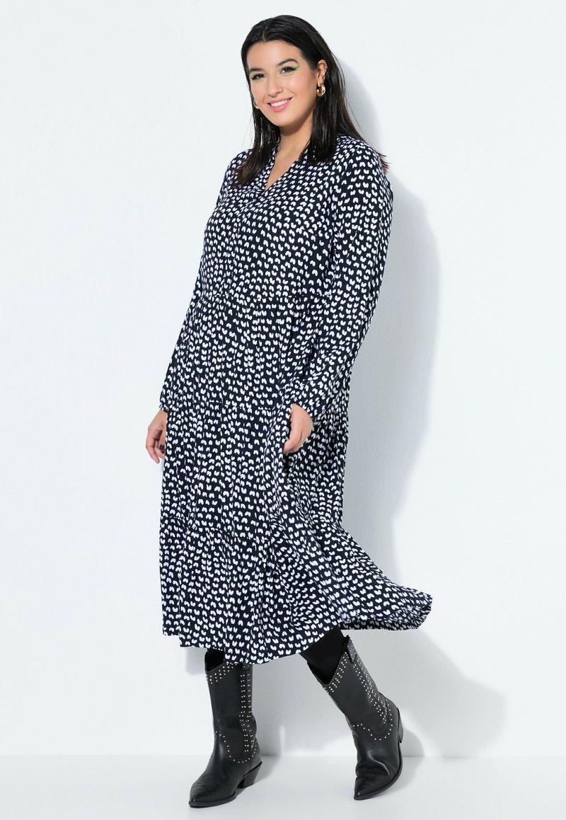 Hymyilevä nainen poseeraa mustavalkoisessa, sydänkuvioisessa pitkässä mekossa mustien leggingsien ja niititettyjen mustien saappaiden kanssa valkoista taustaa vasten.