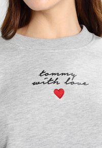 Gros plan d'un sweat-shirt gris avec le texte « tommy with love » et un petit cœur rouge brodé sous le texte sur la poitrine.