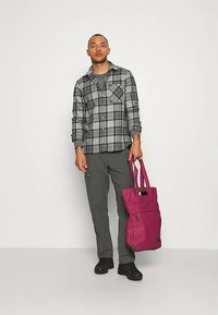 Άνδρας που φορά γκρι πουκάμισο με καρό σχέδιο και σκούρο παντελόνι, κρατώντας μια μεγάλη ροζ τσάντα ώμου, στέκεται μπροστά σε ένα απλό φωτεινό φόντο.