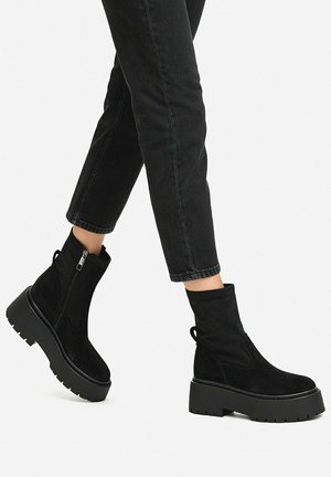 Zwarte suède plateau enkellaarsjes met zijritsen, gedragen met zwarte cropped jeans op een lichte achtergrond.
