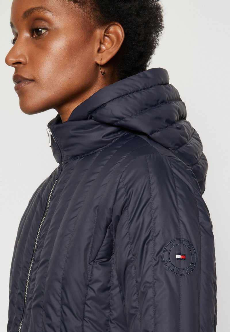 Kobieta z krótkimi włosami ubrana w granatową pikowaną kurtkę z kapturem zapinaną na zamek, z naszywką logo Tommy Hilfiger na rękawie.