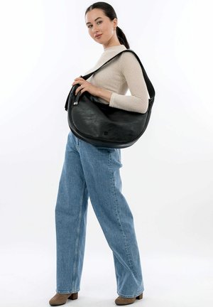 Borsa a spalla in pelle nera con forma curvata, texture liscia e tracolla regolabile, tenuta da una persona che indossa un maglione beige e jeans blu.