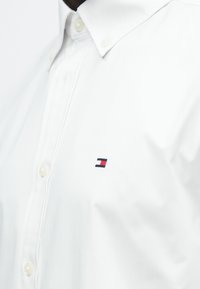 Gros plan sur une chemise blanche boutonnée montrant la zone de la poitrine avec un petit logo brodé en rouge, blanc et bleu marine sur le tissu.